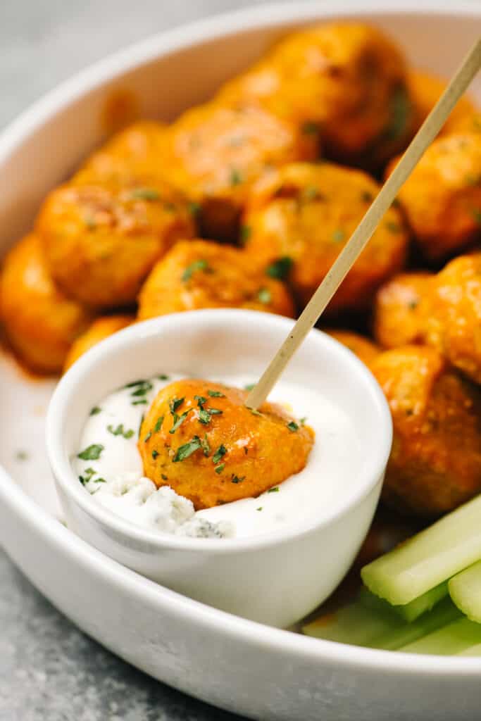 A buffalo chicken meatball on a toothpick being dipped into a small bowl of blue cheese dressing.