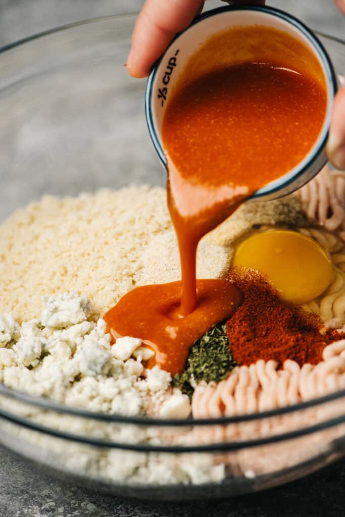 Pouring buffalo sauce into a large bowl with ground chicken, breadcrumbs, blue cheese crumbles, an egg, and seasonings.