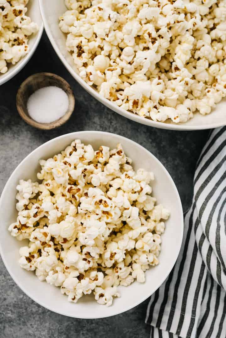 Several bowls of stovetop popcorn on a concrete background with a small dish of salt and a striped linen napkin.