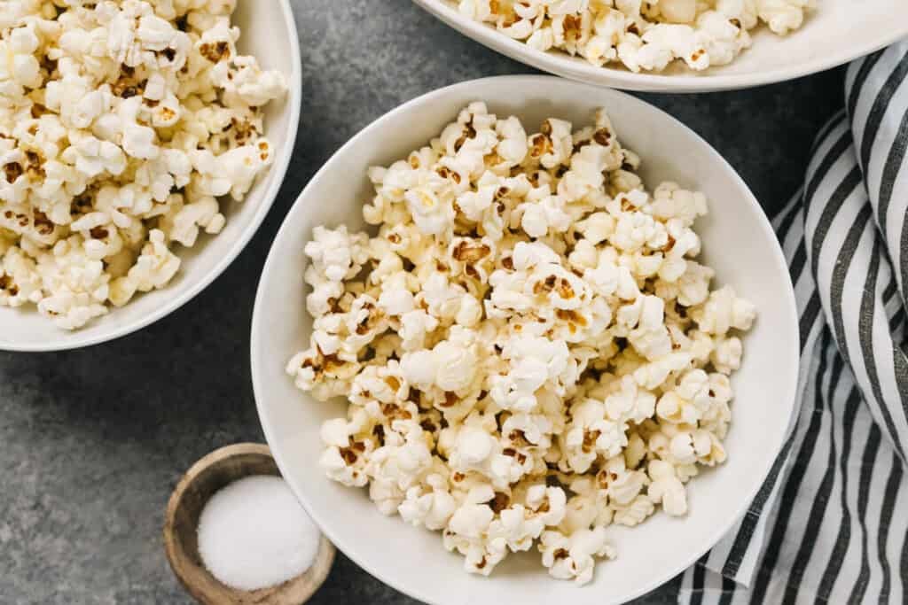 Several bowls of stovetop popcorn on a concrete background with a small dish of salt and a striped linen napkin.