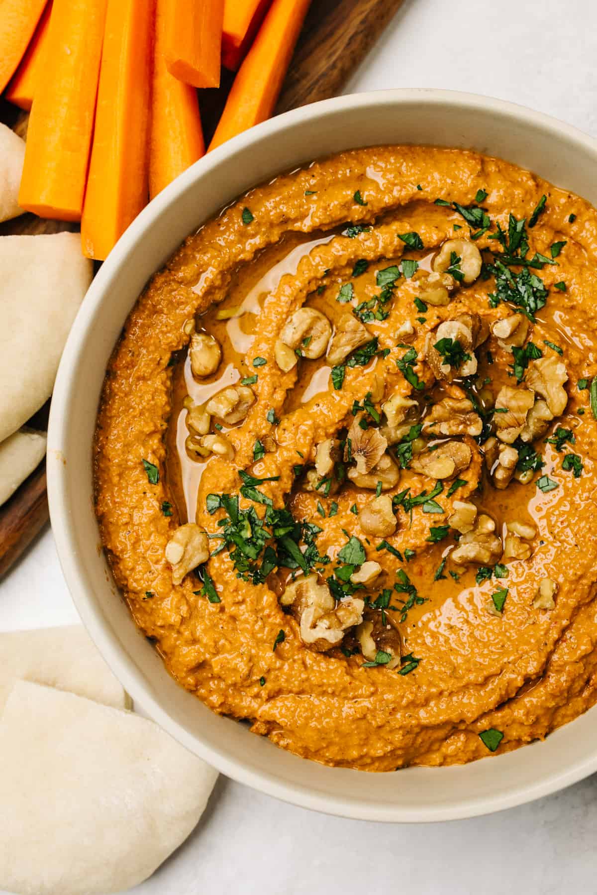Muhammara dip in a low tan bowl, surrounded by pita bread and sliced raw carrots.