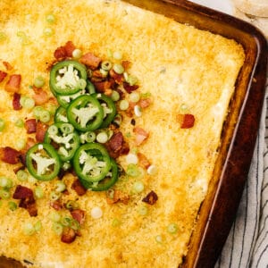 Jalapeno popper dip in a baking dish garnished with bacon, green onions, and sliced jalapeno peppers.